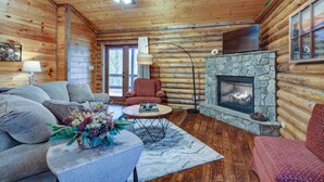 L’intérieur d’une cabane en rondins est agrémenté d’une cheminée en pierre, de planchers en bois, d’un ventilateur de plafond et d’un coin salon confortable avec un canapé et des fauteuils.