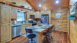 Une cuisine rustique aménagée dans une cabane en rondins, avec des murs en bois, un îlot central doté de tabourets de bar, des appareils électroménagers en acier inoxydable et une horloge au mur.