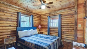 Une chambre à coucher située dans une cabane en rondins, avec un lit, une table de chevet, une lampe et un ventilateur de plafond.