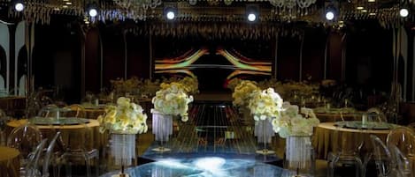 A grand ballroom with a central stage, round tables with chairs, and floral centerpieces.