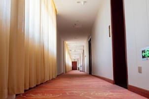 Un pasillo de hotel con alfombra roja, cortinas a la izquierda y puertas a la derecha.