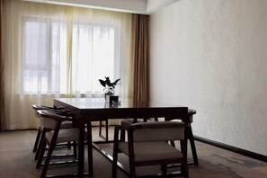 Un salón de conferencias con una larga mesa y seis sillas, ventanas amplias con cortinas translúcidas y un jarrón con plantas sobre la mesa.