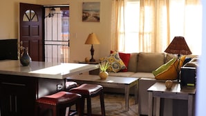 Un soggiorno con divano, tavolino da caffè e sgabelli da bar. Si nota una porta con cancello, una ventola a soffitto e una lampada.