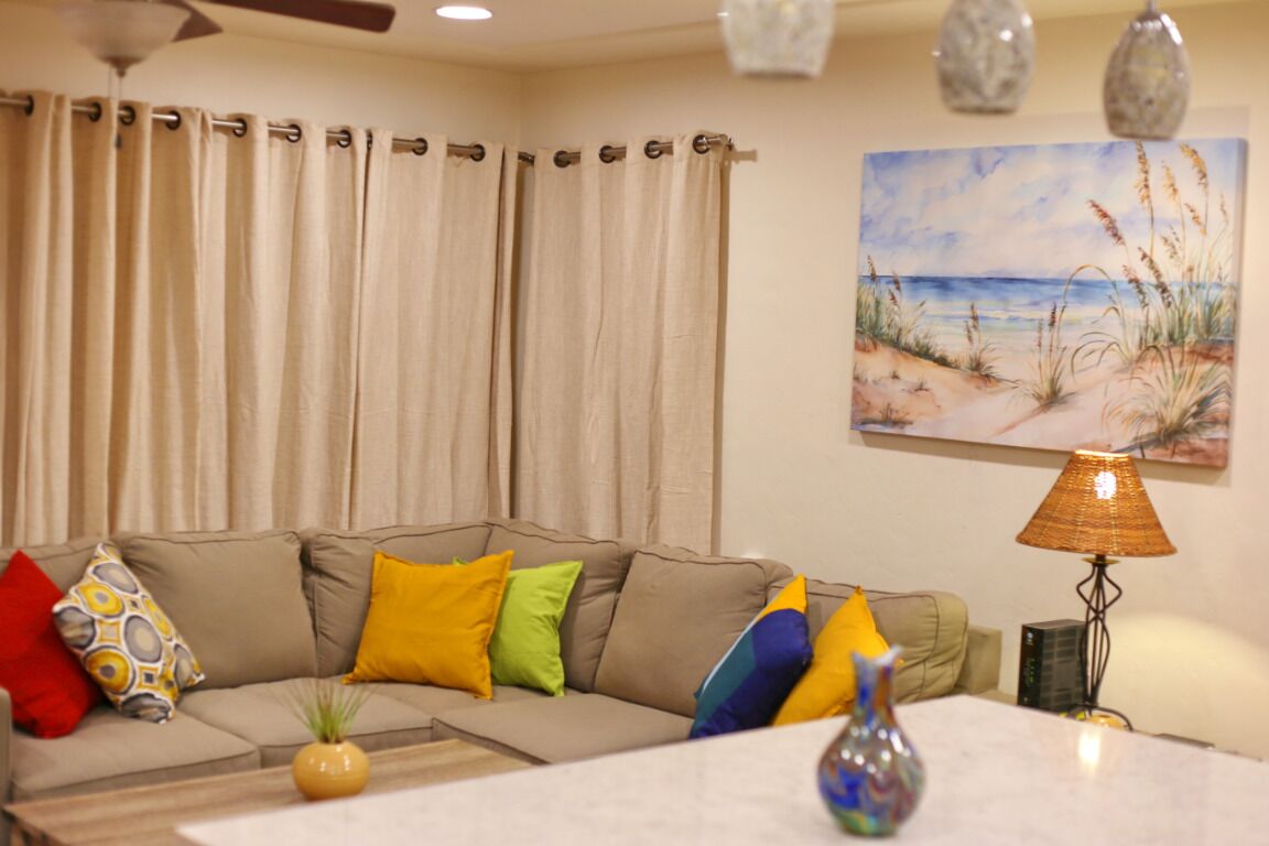 A living room with a beige sofa, colorful cushions, a painting of a beach, and a table lamp.