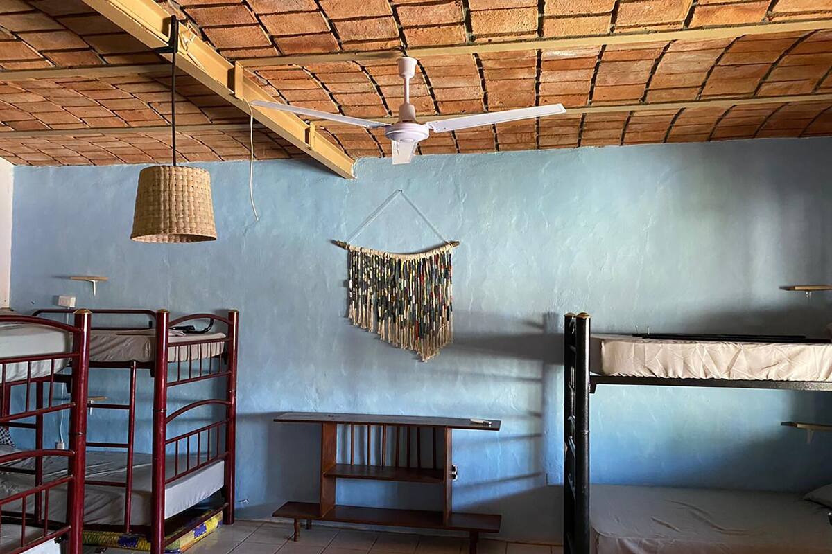 Une chambre de dortoir avec des lits superposés, un ventilateur de plafond, une lampe en fibres tressées et une décoration murale en macramé.