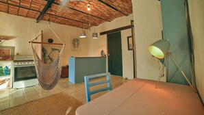 A cozy living room with a hammock, a dining table, a lamp, and a kitchen area with an oven.