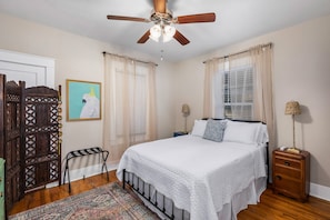 Un dormitorio con cama, ventilador de techo, mesita de noche, ventana con cortinas, puerta y una pared con un cuadro enmarcado.