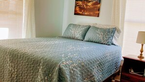 A bedroom with a bed, a nightstand, a lamp, a painting, and a window with blinds.