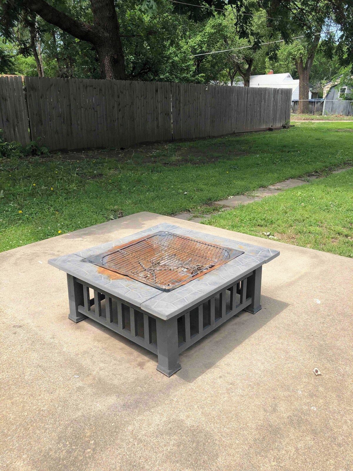 Ein quadratisches Außenfeuer mit einem Metallrost, umgeben von einer Betonbasis, platziert in einer Rasenfläche mit einem Holzzaun im Hintergrund.