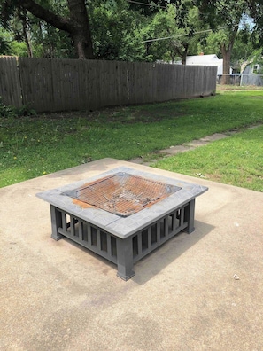 Una chimenea exterior cuadrada con rejilla metálica, rodeada por una base de concreto, ubicada en un área con césped y una valla de madera al fondo.