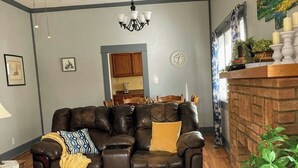 A living room with a leather sofa, a patterned rug, a ceiling fan, a chandelier, and a brick fireplace.