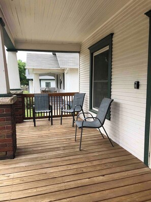A covered porch with a wooden deck, a brick fireplace, and two chairs. - Stay Awhile Your Comfortable Stay (Hutchinson)