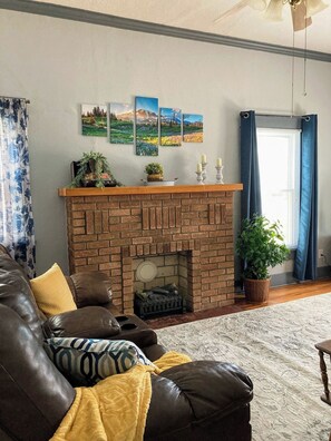 Una sala de estar con chimenea de ladrillo, un sofá de cuero y un amplio ventanal con cortinas azules.