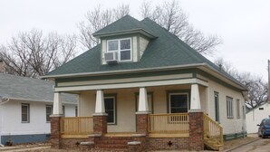 A two-story house with a green roof, a front porch with a wooden railing, and a brick foundation.