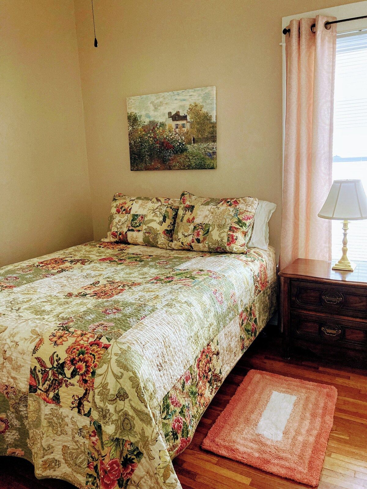 A bedroom with a floral bedspread, a painting on the wall, a bedside table with a lamp, and a wooden floor.