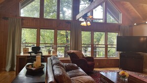 A spacious living room with large windows, a leather sofa, a dining table, and a television. - Lake Front, Beautiful views, Gentle slope to Lake, Hot tub, Close to Downtown. (Morganton)