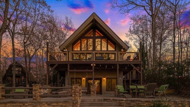 A two-story log cabin with a prominent gable roof, a stone foundation, and a front porch with outdoor seating. - Lake Front, Beautiful views, Gentle slope to Lake, Hot tub, Close to Downtown. (Morganton)