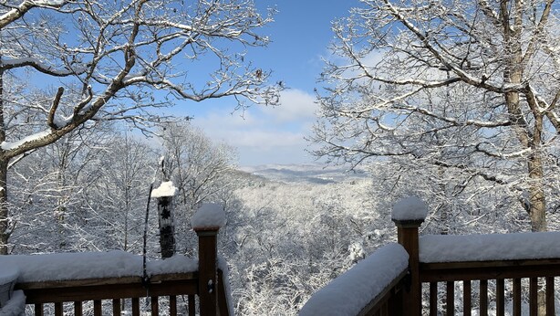 Eine Holzterrasse mit schneebedeckten Geländern und Blick auf verschneite Bäume und Berge im Hintergrund.
