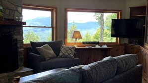 Ett mysigt vardagsrum med en öppen spis i sten, en soffa, ett soffbord och en tv. Det finns två fönster med utsikt över bergen.