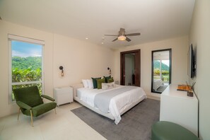 A modern bedroom with a large bed, a green armchair, a desk, and a view of greenery through the window.