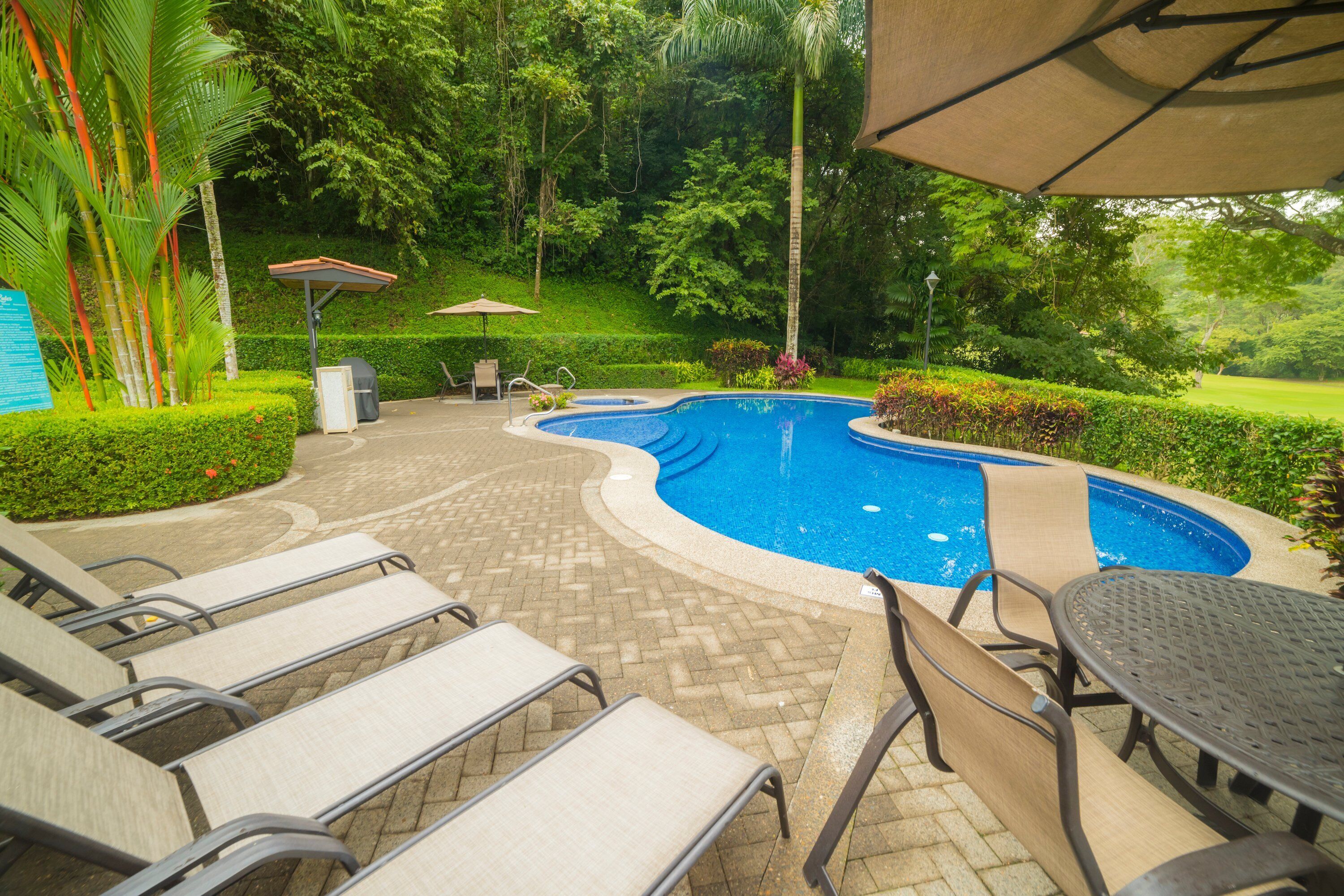 A pool area with lounge chairs and a table, surrounded by greenery and a well-maintained lawn.