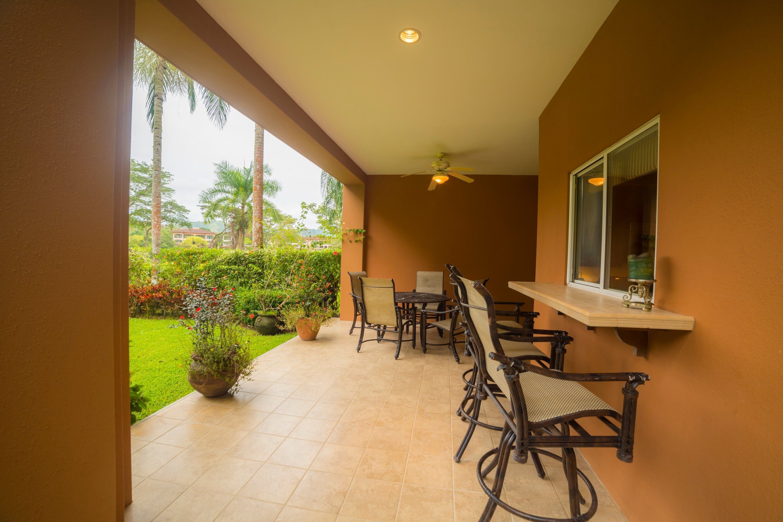 A covered patio with a table and chairs, a ceiling fan, and a window overlooking a garden with palm trees and a building in the background.