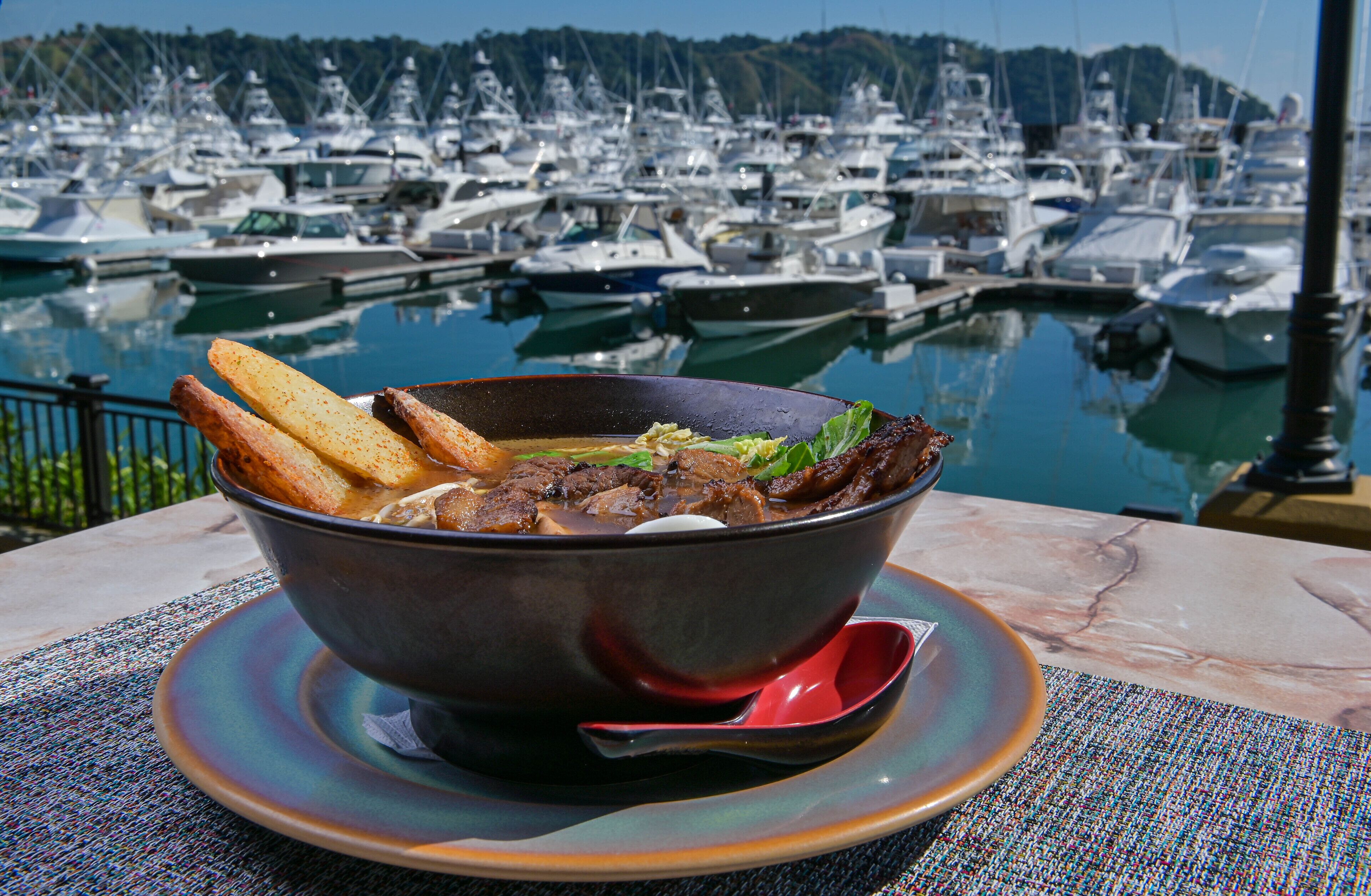 Eine Schüssel Suppe mit Brot auf einem Tisch, im Hintergrund Blick auf eine Marina.