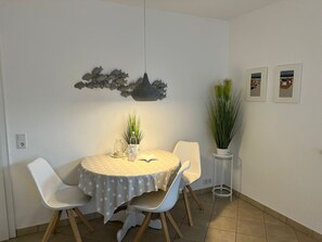 Un área de comedor con una mesa redonda cubierta con un mantel estampado de estrellas, rodeada de sillas blancas. Hay una lámpara colgante sobre la mesa, una decoración de peces en la pared y dos plantas en macetas.