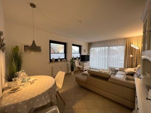 Una sala moderna con un sofá, una mesa de comedor con sillas, un televisor y un ventanal con cortinas.