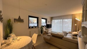 A modern living room with a sofa, a dining table with chairs, a TV, and a large window with curtains. - Margeritenhaus - Relaxed island life: Cozy vacation apartment in Westerland (Sylt)