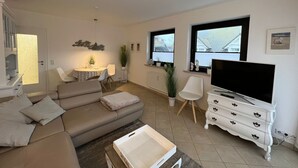 A modern living room with a sofa, a dining table, a TV, and a white dresser. - Margeritenhaus - Relaxed island life: Cozy vacation apartment in Westerland (Sylt)