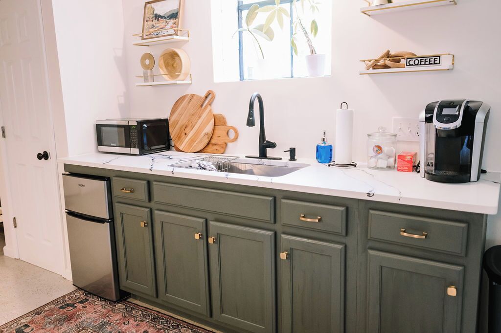 Una cocina con encimera blanca, un lavabo, microondas, cafetera y armarios verdes.