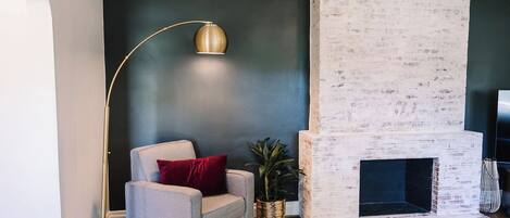 A living room with a white brick fireplace, a grey armchair with a red pillow, a floor lamp, and a potted plant.