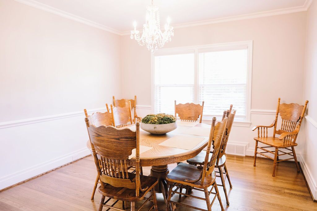 A dining room with a round table surrounded by wooden chairs, a chandelier, and a window with blinds.