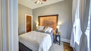 A bedroom with a bed, a wooden door, a brick accent wall, a ceiling fan, and a window with curtains. - Historic Downtown Hawley Loft (Hawley)