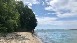 Une plage rocheuse où l’on voit une personne debout près de l’eau, des arbres en arrière-plan et un ciel dégagé.