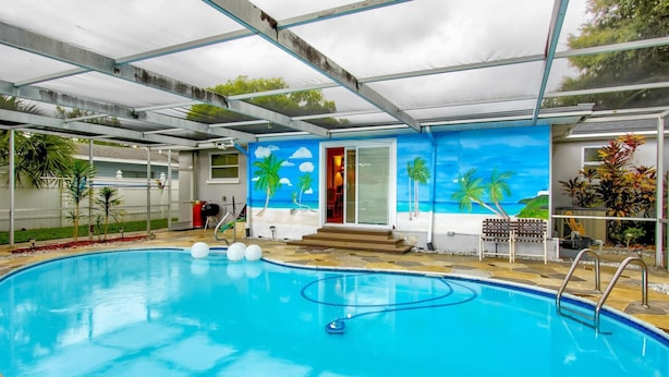 Ein Pool mit klarem, blauem Wasser, ein überdachter Bereich mit Metallveranda und ein Wandgemälde mit Strandmotiv.