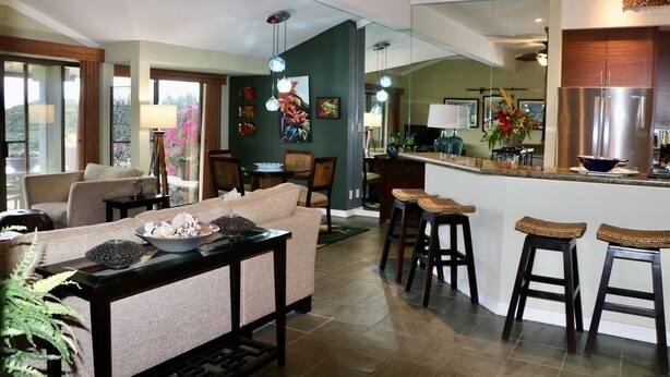 Una sala moderna con sofá, mesa de centro y taburetes de bar. Se ve una cocina con refrigerador y mostrador de bar al fondo.