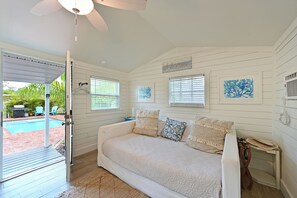 A room with a white wooden wall, a white sofa with patterned cushions, a ceiling fan, and a view of a swimming pool outside. - Historic 1930s Cottage + Heated Pool + 12mi to beaches & 1 mi to downtown (Palmetto)