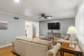 A living room with a beige sofa, a wooden coffee table, a TV mounted on the wall, and a ceiling fan.