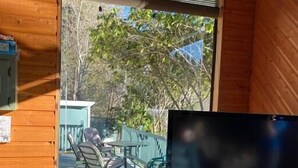 Una cabaña de madera con un televisor sobre un mueble de madera, y un ventanal corredero que da a una terraza exterior con sillas y una mesa.