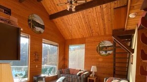 Interior acogedor de una cabaña con techo alto, paredes de madera, chimenea, un sofá, un sillón y un televisor de pantalla plana.