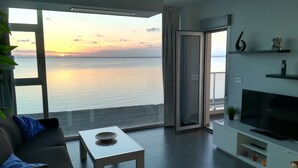 Een moderne woonkamer met een grote televisie, een bank, een salontafel en uitzicht op zee bij zonsondergang.