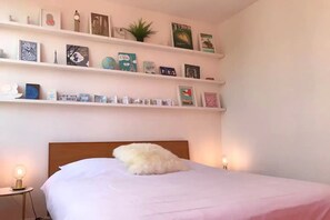 A bedroom with a bed, a headboard, and a shelf above it containing books and decorative items.