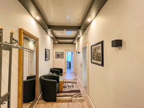 Um corredor com piso de madeira, cadeiras pretas, tapete estampado, quadros nas paredes e teto com iluminação embutida.