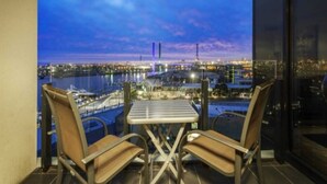 A balcony with two chairs and a table, overlooking a cityscape at dusk with a bridge and industrial structures in the background.