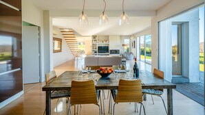 Une salle à manger spacieuse avec une longue table, des chaises et un panier de fruits. Des luminaires suspendus sont placés au-dessus de la table, et on aperçoit un coin cuisine à l’arrière-plan.