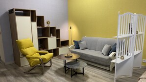 Una sala de estar con una pared amarilla, un sofá gris, un sillón amarillo, una estantería de madera y una puerta de seguridad blanca para bebés.