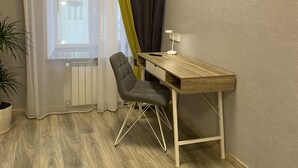 Una habitación pequeña y ordenada con un escritorio de madera, una silla, una planta en maceta y un ventanal con cortinas de gasa y un toldo oscuro.