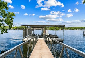 A wooden dock leading to a covered boat launch area with a view of a calm lake and distant buildings.